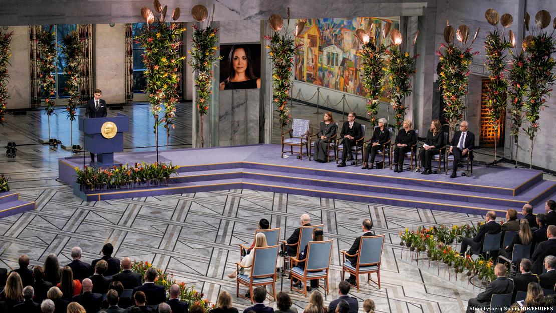 Ceremonia de entrega del Nobel de la Paz a María Corina Machado en Oslo, con una foto de ella en el centro del podio. Ceremonia de entrega del Nobel de la Paz a María Corina Machado en Oslo, con una foto de ella en el centro del podio.