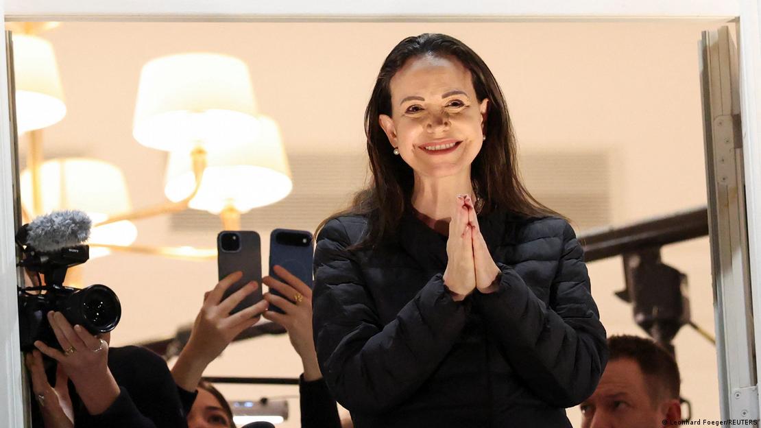 María Corina Machado de mãos postas com pessoas fotografando com celular ao fundo
