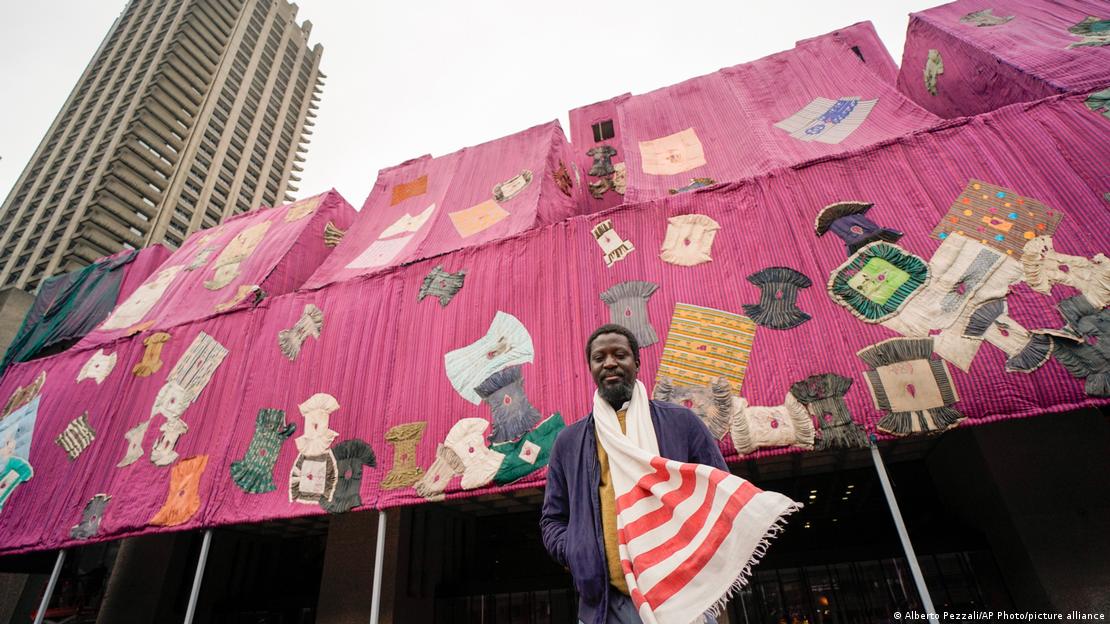 Ο Μαχάμα στέκεται μπροστά από ένα μεγάλο κτίριο που καλύπτεται από ένα μεγάλο ροζ-μωβ ύφασμα που είναι έντονα χρωματισμένο ρόμπες ραμμένες πάνω του