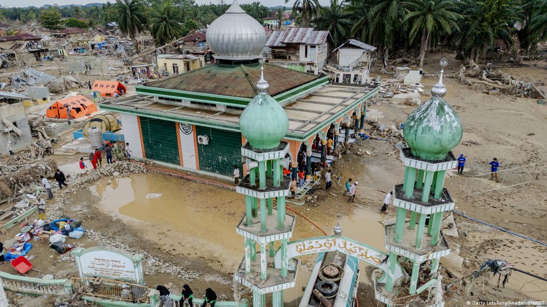Fotoğraf havadan çekildi. Ortada yeşil-beyaz renkte bir cami var. Caminin iki ince minaresi ve ortasında büyük, yuvarlak bir kubbesi bulunuyor. Kubbe ve minareler ayakta, ancak caminin etrafı tamamen çamur ve enkazla çevrili. Fotoğraf havadan çekildi. Ortada yeşil-beyaz renkte bir cami var. Caminin iki ince minaresi ve ortasında büyük, yuvarlak bir kubbesi bulunuyor. Kubbe ve minareler ayakta, ancak caminin etrafı tamamen çamur ve enkazla çevrili.