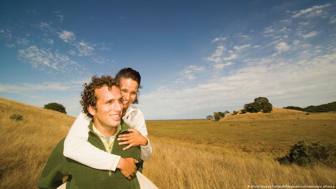 un hombre joven lleva en sus espaldas a una joven mujer, ambos sonríen, en el campo