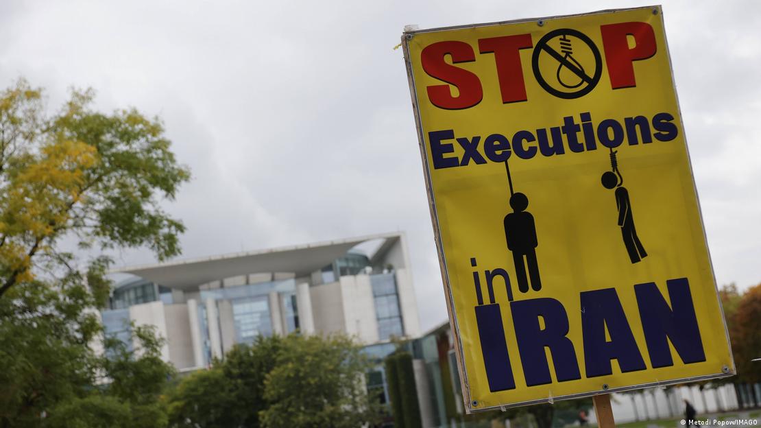 "Stop Executions in Iran" ("Stoppt Hinrichtungen im Iran") steht auf einem Plakat, das eine auf dem Foto nicht zu sehende Person gegenüber vom Kanzleramt in Berlin in die Höhe hält. Zwischen den Wörtern sind zwei Piktogramme von erhängten Menschen zu sehen