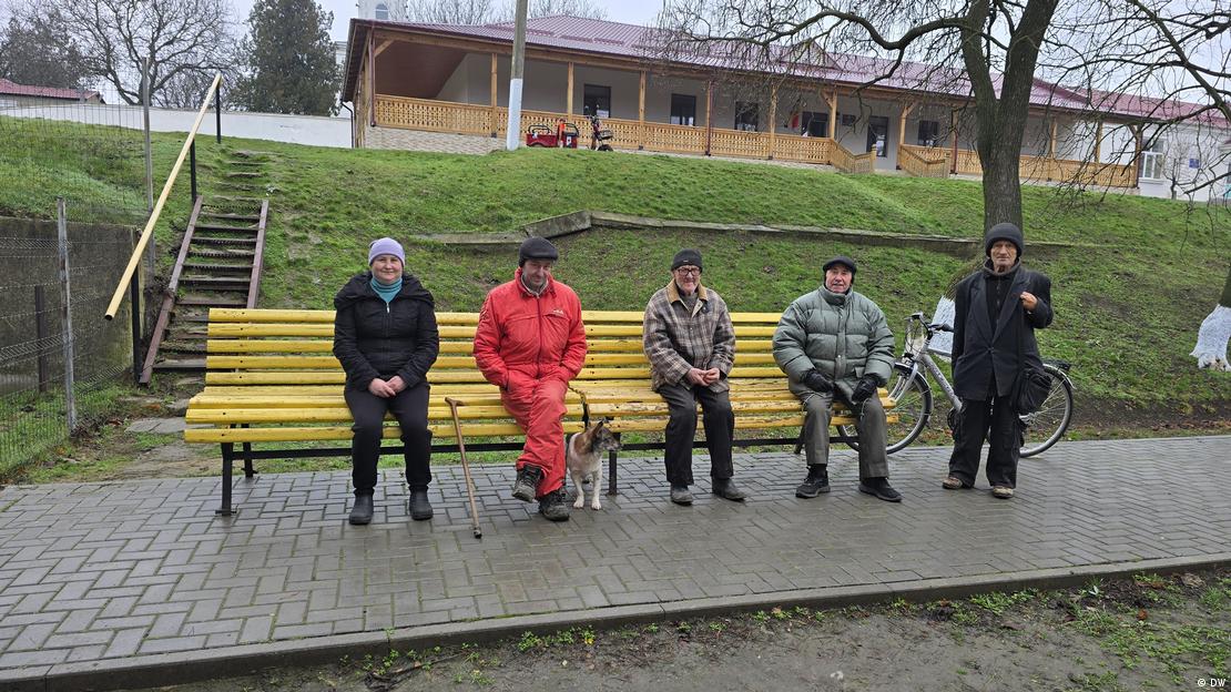 Oameni stau aşezaţi pe o bancă şi aşteaptă să le fie dată o masă la Cantina Socială