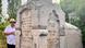 Ein Mann mit weißem Hemd und Hut, der eine Brille trägt, steht neben einem großen, kunstvoll gestalteten Grabstein auf einem Friedhof Ein Mann mit weißem Hemd und Hut, der eine Brille trägt, steht neben einem großen, kunstvoll gestalteten Grabstein auf einem Friedhof