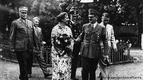 Лидерот на нацистичка Германија, Адолф Хитлер (втор од десно) разговара со жена на црно-бела фотографија. Опкружени се со четворица мажи од кои двајца во воени униформи. 
