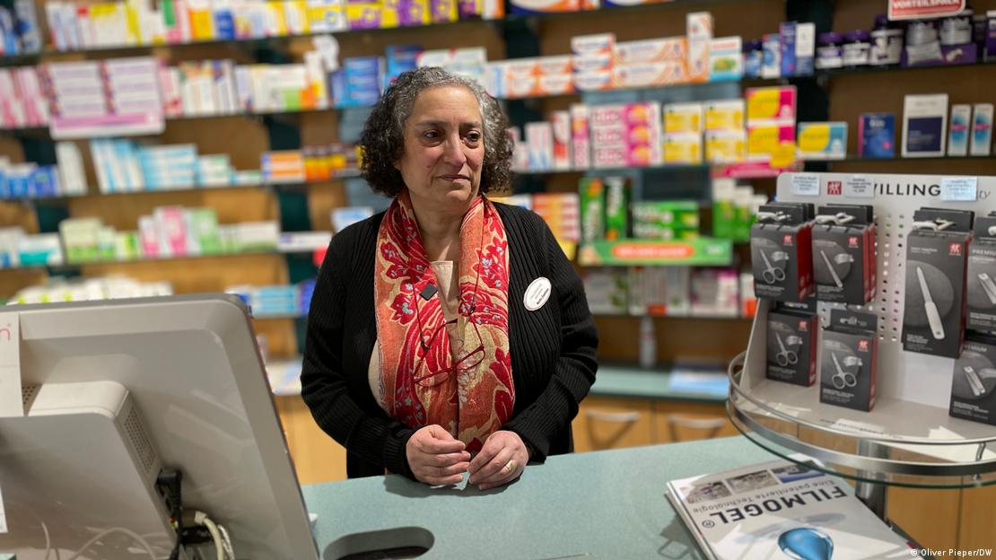 Eine Frau mit einem roten geblümten Schal steht vor einem Regal mit Medikamenten, sie sieht traurig aus