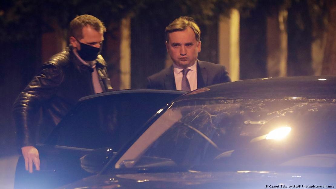 Двајца мажи, едниот со костум, а другиот со темна јакна и маска на лицето стојат покрај автомобил со отворена врата