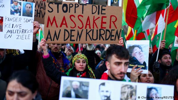 Berlin'deki protesto gösterisine katılan kalabalık görülüyor. Bir kadın eylemcinin elinde "Kürtlere katliamı durdurun" yazılı pankart var
