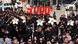 People wearing black march while holding white flowers and a large red sign that reads '5,000' People wearing black march while holding white flowers and a large red sign that reads '5,000'