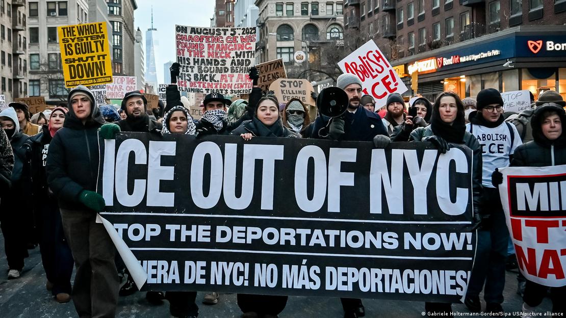 Multidão em passeata na rua com uma grande faixa negra com letras brancas: "ICE out of NYC" e "Stop the deportations now!"