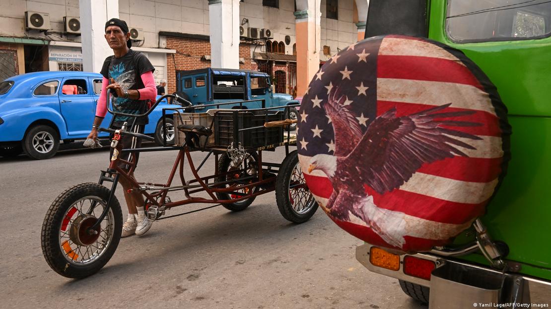 Cubano en un vehículo a pedales con autos estacionados, uno de los cuales lleva la rueda de repuesto cubierta con una funda con la imagen de la bandera de Estados Unidos y un águila.
