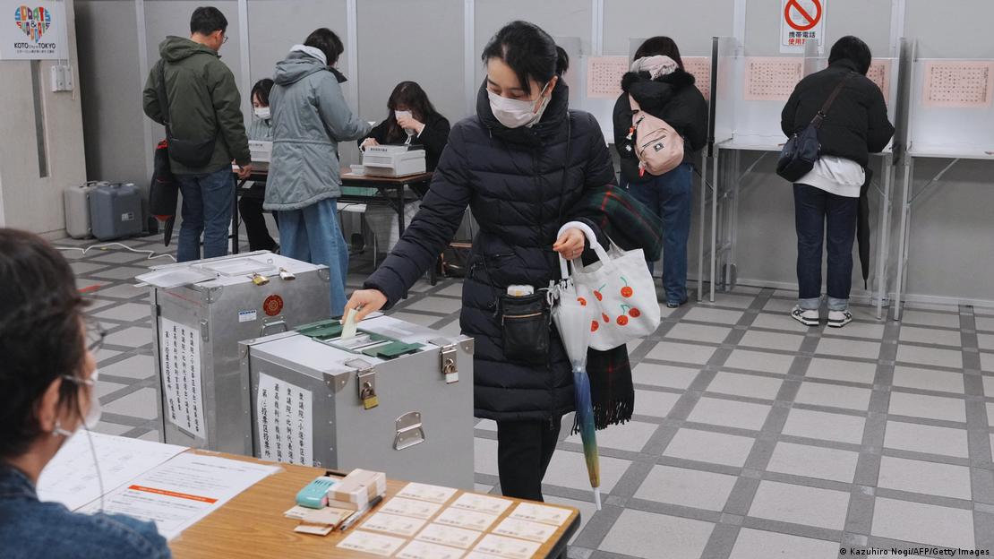 La mujer, con abrigo, tapabocas y paraguas, mete su voto en la urna.