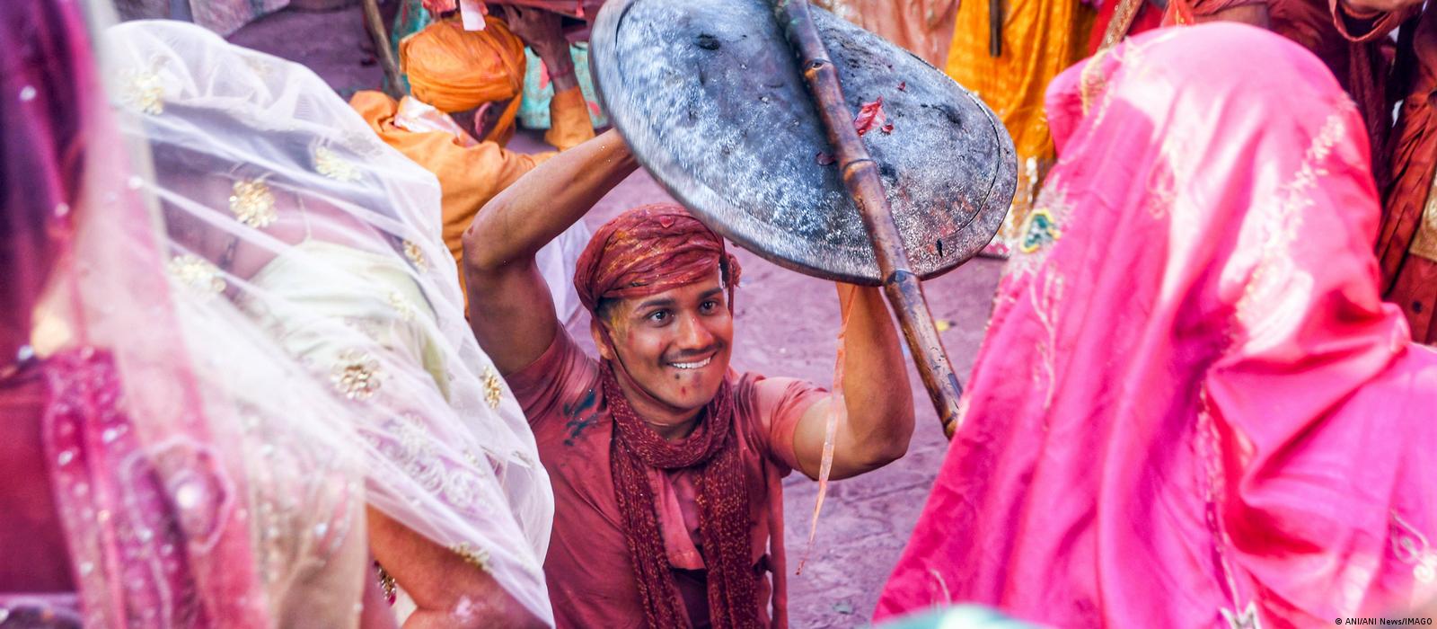 A smiling man kneels on the ground and holds a round shield above his head, which a woman, seen only from behind, hits with a bamboo stick. Colorfully dressed people can be seen around the two.