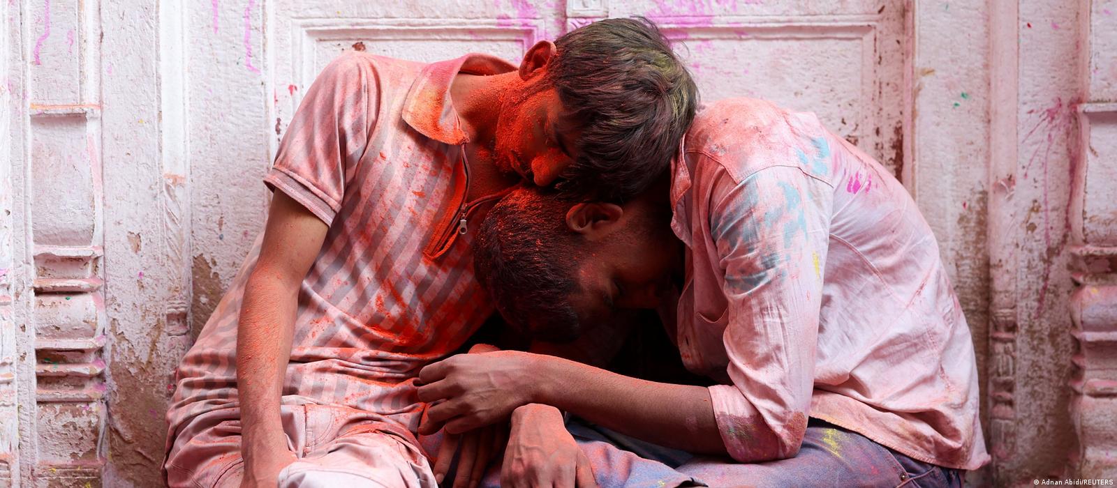 Two young men are sitting in front of a paint-splattered wall; they have their heads leaning against each other and their eyes closed. 