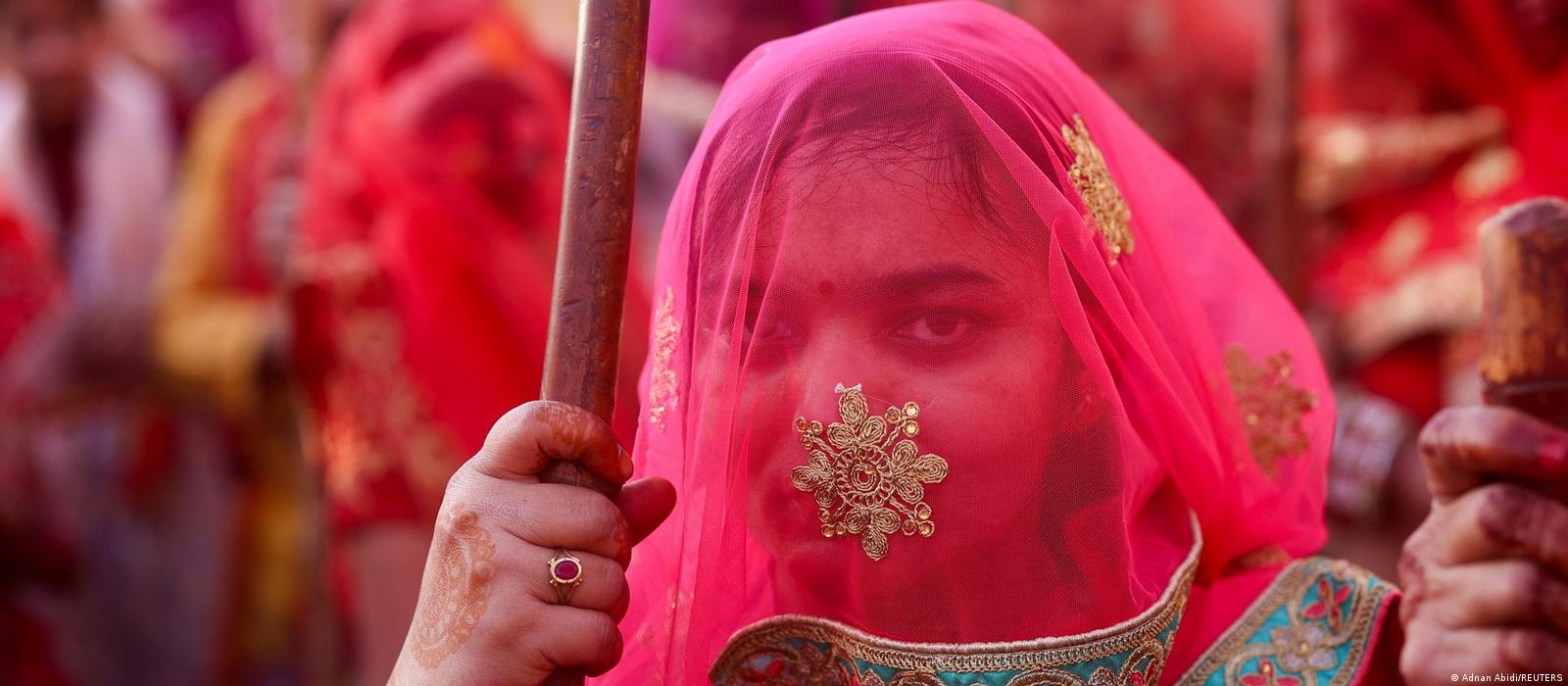 A woman whose face is covered by a transparent pink veil with gold appliqués looks into the camera. She is holding a wooden stick in her hand.