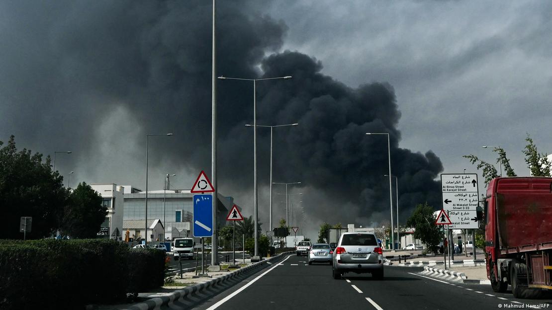 Im Zentrum des Bildes steht in deutlicher Entfernung von der Kamera eine gewaltige schwarze Rauchsäule, die zurückzuführen ist auf einen iranischen Angriff auf das Industriegebiet von Doha/Katar. Der Fotograf blickt wohl aus einem fahrenden Auto. Vor ihm bewegen sich andere Fahrzeuge auf einer mehrspurigen Straße auf die Rauchsäule zu.