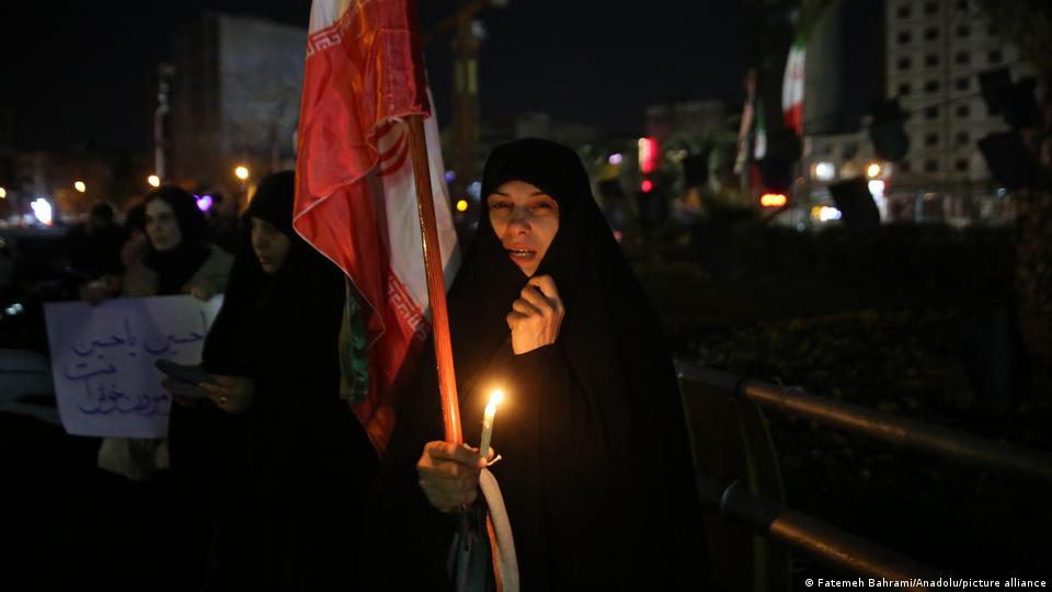 Con una vela y una bandera iraní, una mujer vestida de negro y cubierta con el velo islámico llora. Al fondo, otros manifestantes que no se distinguen.