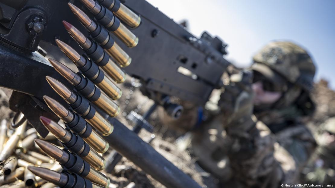 Un soldado apunta con su ametralladora. En primer plano se ve una cinta con balas.