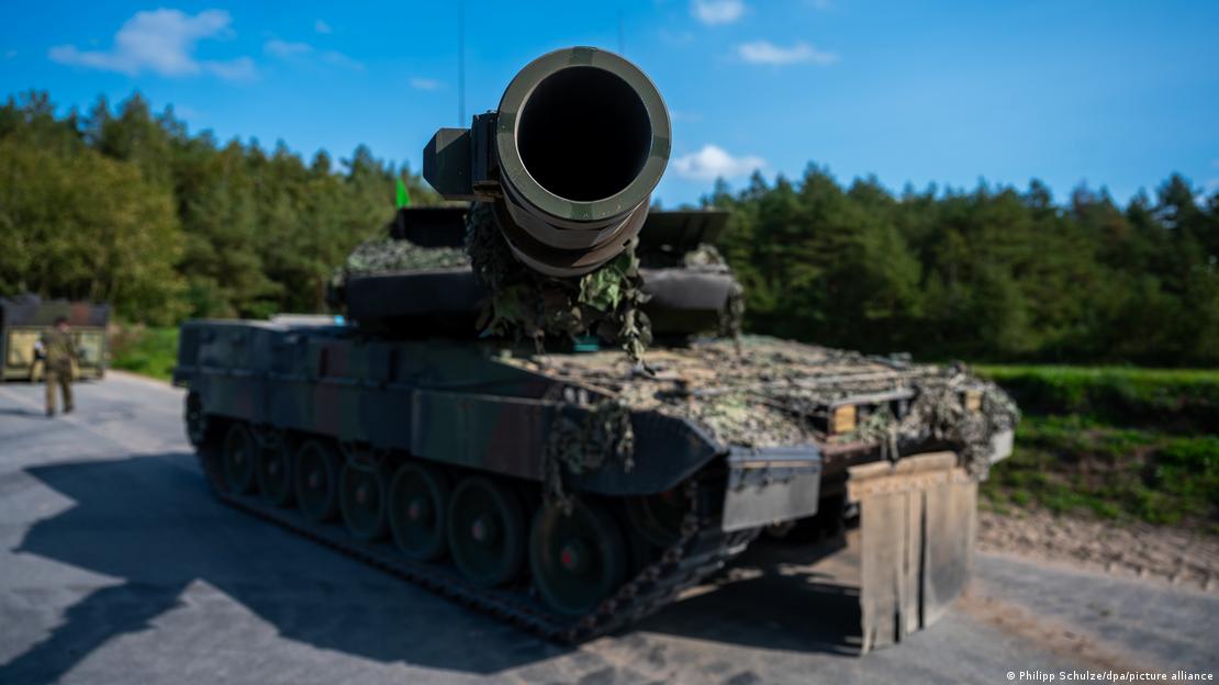Man blickt in das Geschützrohr des Kampfpanzer Leopard 2 A7V, der auf einem Übungsplatz der Bundeswehr steht. Der Hintergrund ist bewaldet, man sieht am linken Bildrand unscharf einen Soldaten. 