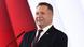 Przemyslaw Czarnek, a middle-aged man with short hair, stands at a microphone in front of a red and white Polish flag, smiling Przemyslaw Czarnek, a middle-aged man with short hair, stands at a microphone in front of a red and white Polish flag, smiling