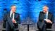 Two middle-aged men seated on a debate stage, both wearing dark blue suits, white shirts and dark blue ties. The one on the left is wearing glasses. They are looking at each other over a small round table holding water glasses and a carafe Two middle-aged men seated on a debate stage, both wearing dark blue suits, white shirts and dark blue ties. The one on the left is wearing glasses. They are looking at each other over a small round table holding water glasses and a carafe