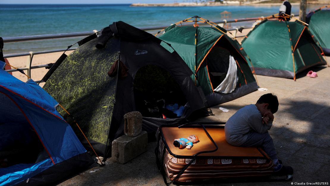 Sahil kenarındaki çadırların önünde bir bavulun üstüne oturmuş küçük bir erkek çocuğu