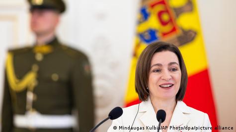 Një grua pas një mikrofoni (Maia Sandu), me një ushtar dhe një flamur të paqartë në sfond.