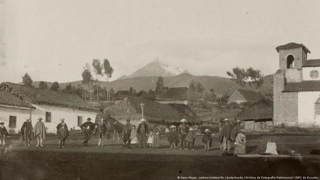 Imagen en blanco y negro: personas en una aldea. Un volcán de fondo.