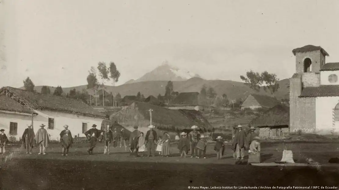 Imagen en blanco y negro: personas en una aldea. Un volcán de fondo.