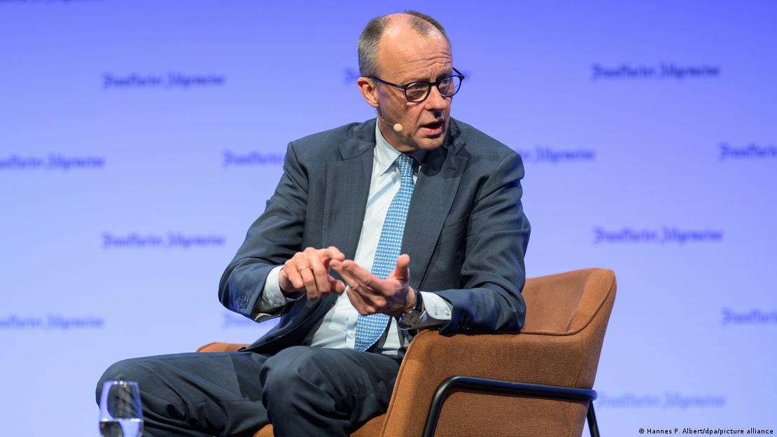 Friedrich Merz u odijelu sjedi u smeđem naslonjaču ispred plave pozadine