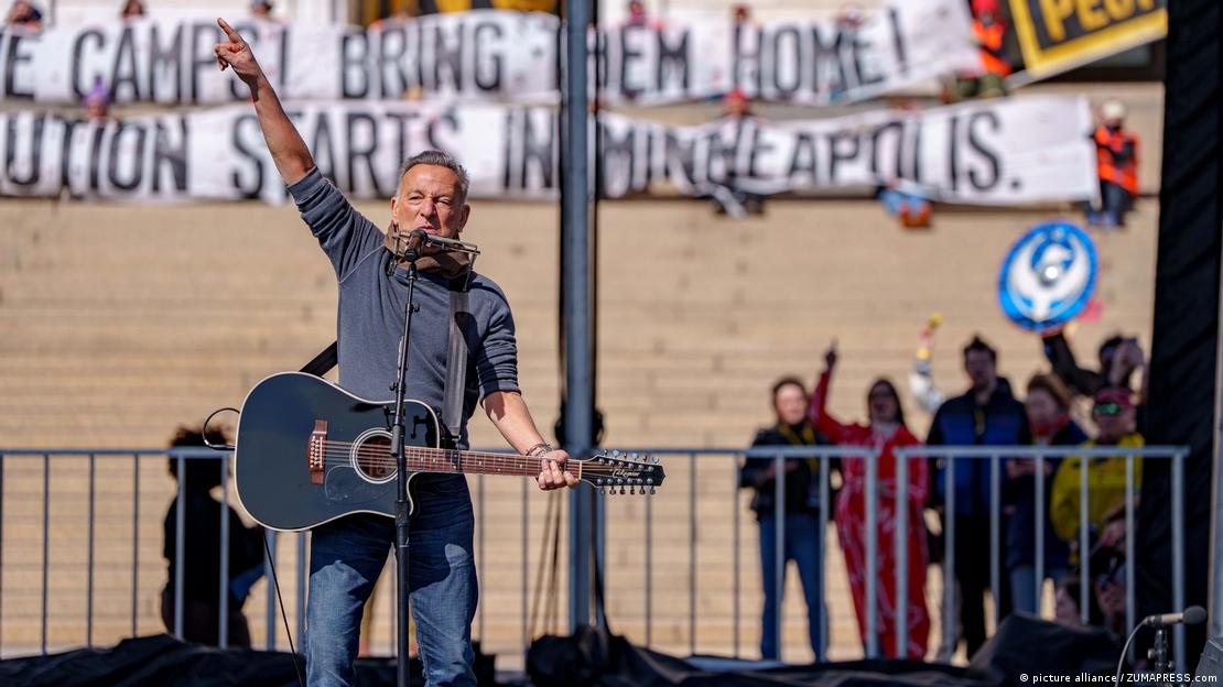 O cantor americano Bruce Springsteen ergue os braços e acena para o público enquanto canta e toca violão