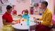 An exemplary family (father, mother, and child) is sitting together at a small round table in a child-friendly setting made entirely of plastic. On the table are plastic plates, plastic cups, and plastic food. In the background, the apartment is also furnished entirely with plastic. An exemplary family (father, mother, and child) is sitting together at a small round table in a child-friendly setting made entirely of plastic. On the table are plastic plates, plastic cups, and plastic food. In the background, the apartment is also furnished entirely with plastic.