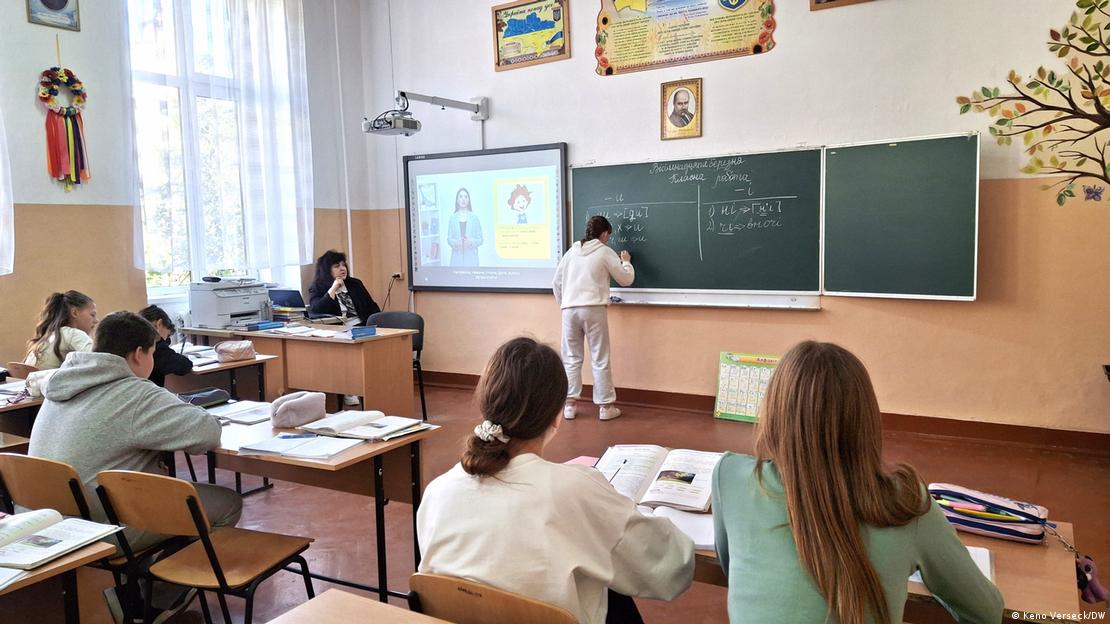 Ein Klassenraum mit Blick auf die Tafel, vor der ein Mädchen steht und etwas an die Tafel schreibt. Außerdem sind einige Schüler und die Lehrerin zu sehen