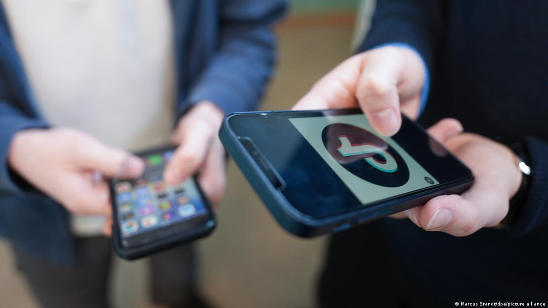 İki kişinin elinde akıllı telefonlar bulunuyor. Telefonlardan birinin ekranında TikTok logosu yer alıyor; diğer telefon ekrana dönük şekilde tutuluyor. Görsel, mobil iletişim ve dijital platformlara erişimi sembolize ediyor