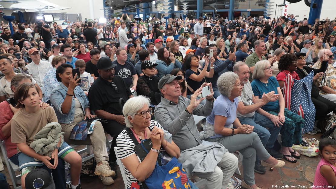 Multidão sentada em cadeiras, com várias pessoas segurando celulares, enquanto assistem à chegada da Artemis 2