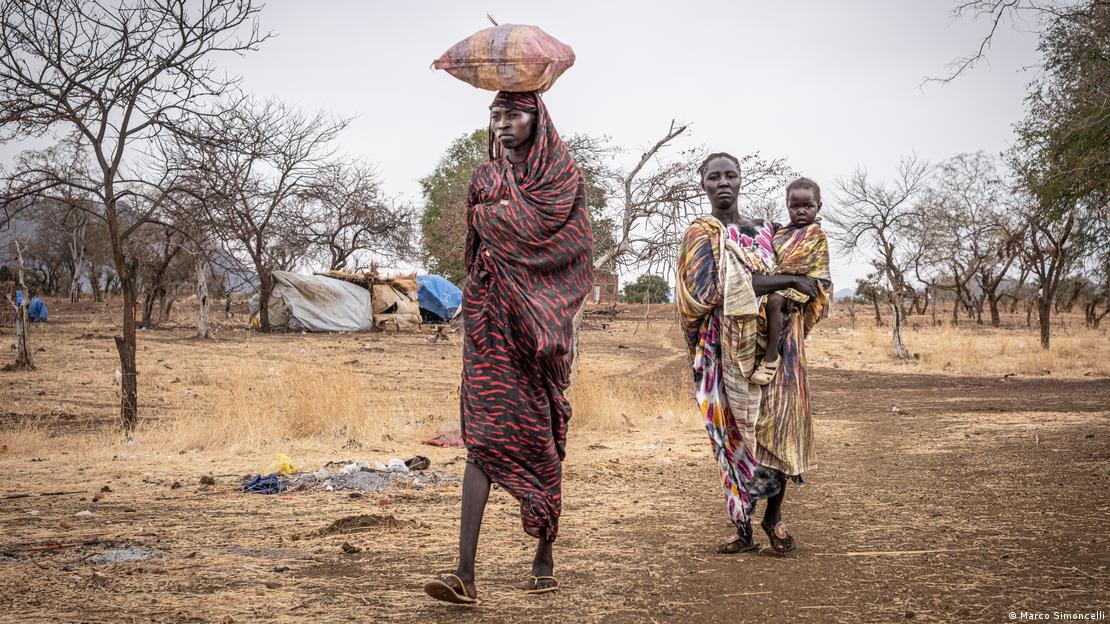 Uma mulher segue na frente equilibrando um saco de grãos na cabela, outra segue atrás carregando um bebê 