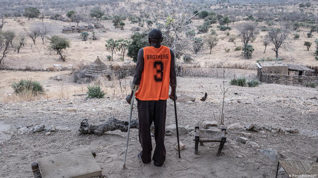 Hassan Koko apoiado em muletas, em região de vegetação árida 