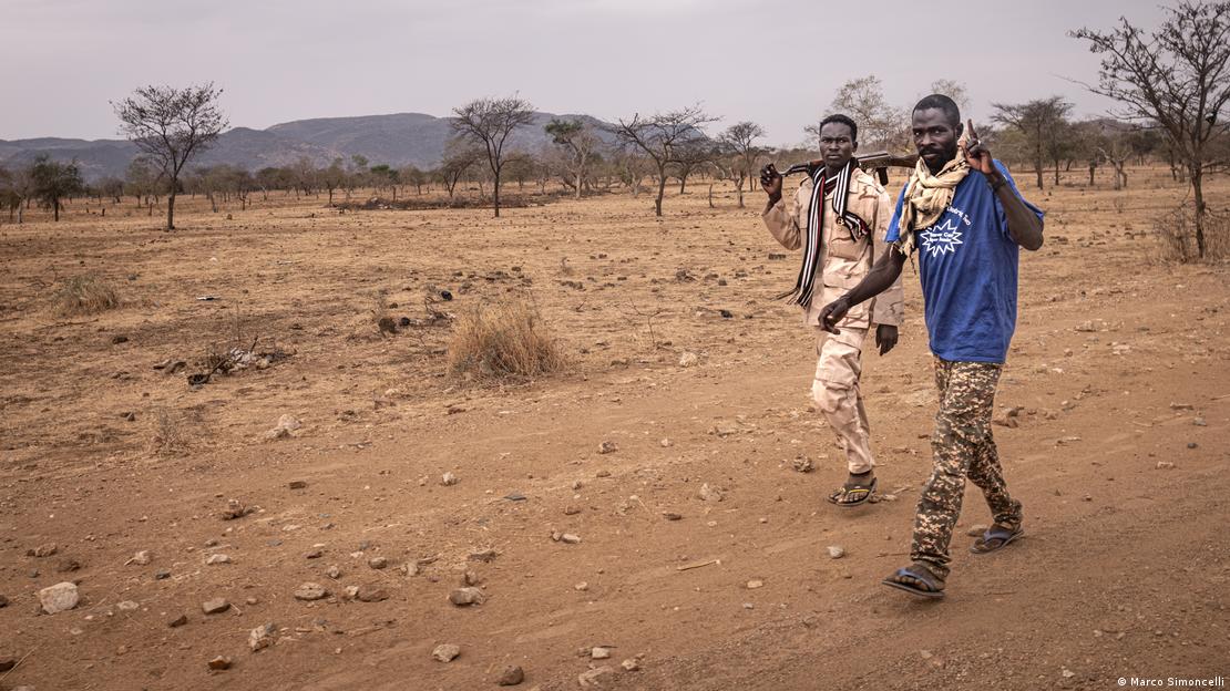 Dois homens com armas nos ombros caminham em estrada de terra