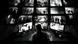 Black-and-white stylized depiction of a surveillance control room: a person sits at a desk facing multiple monitors displaying different camera views of indoor and outdoor spaces with people and architectural structures. Black-and-white stylized depiction of a surveillance control room: a person sits at a desk facing multiple monitors displaying different camera views of indoor and outdoor spaces with people and architectural structures.