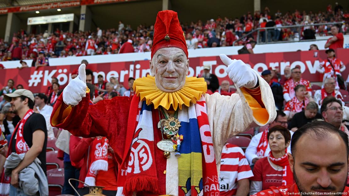 Mainz-Bayern Münih maçında tribünde poz veren bir Mainz taraftarı. Taraftarın yüzünde makyaj, üzerinde ise kırmızı-beyaz ağırlıklı karnaval kostümü ve takım atkısı var