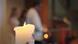 Lit candle in the foreground of a church interior, with blurred seated people on benches in the background, calm atmosphere, symbolic light during a church service or vigil. Lit candle in the foreground of a church interior, with blurred seated people on benches in the background, calm atmosphere, symbolic light during a church service or vigil.