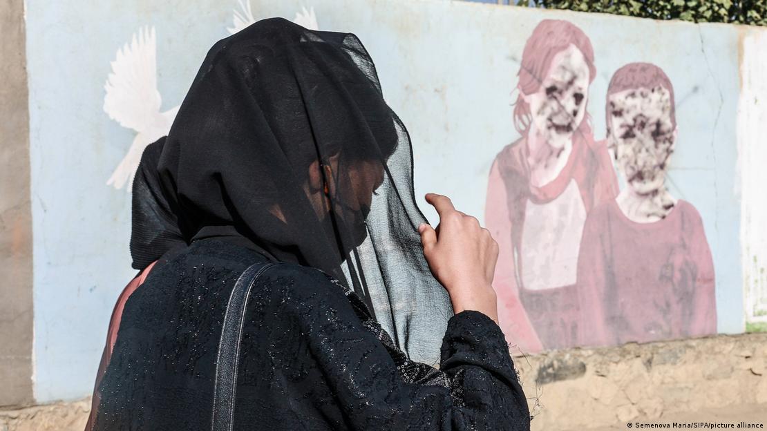 Başı kapalı, yüzü tülden bir örtü ile örtülü bir kadın, üstünde yüzleri tanınmaz hale getirilmiş bir kız, bir erkek çocuk resminin olduğu duvarın önünden geçiyor