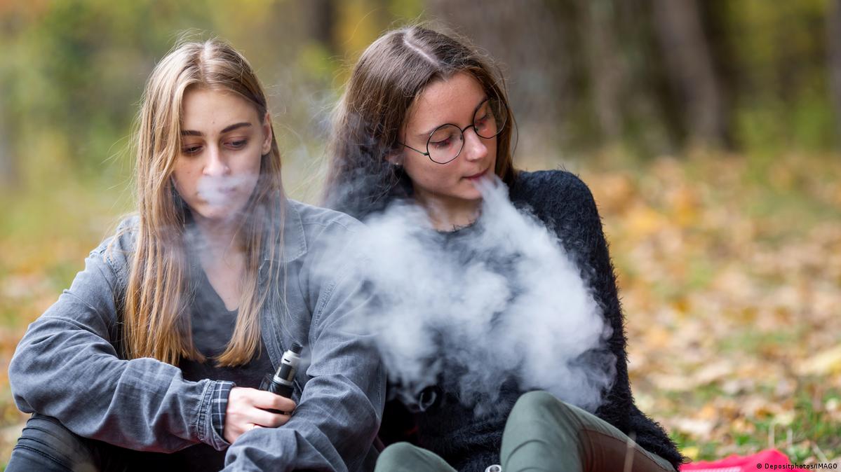 Teenage girls in casual clothes vape electronic cigarettes in a park.