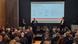 Uczestnicy konferencji sieszą na krzesłach, w tle perzentacja na ekranie i trzech panelistów Uczestnicy konferencji sieszą na krzesłach, w tle perzentacja na ekranie i trzech panelistów