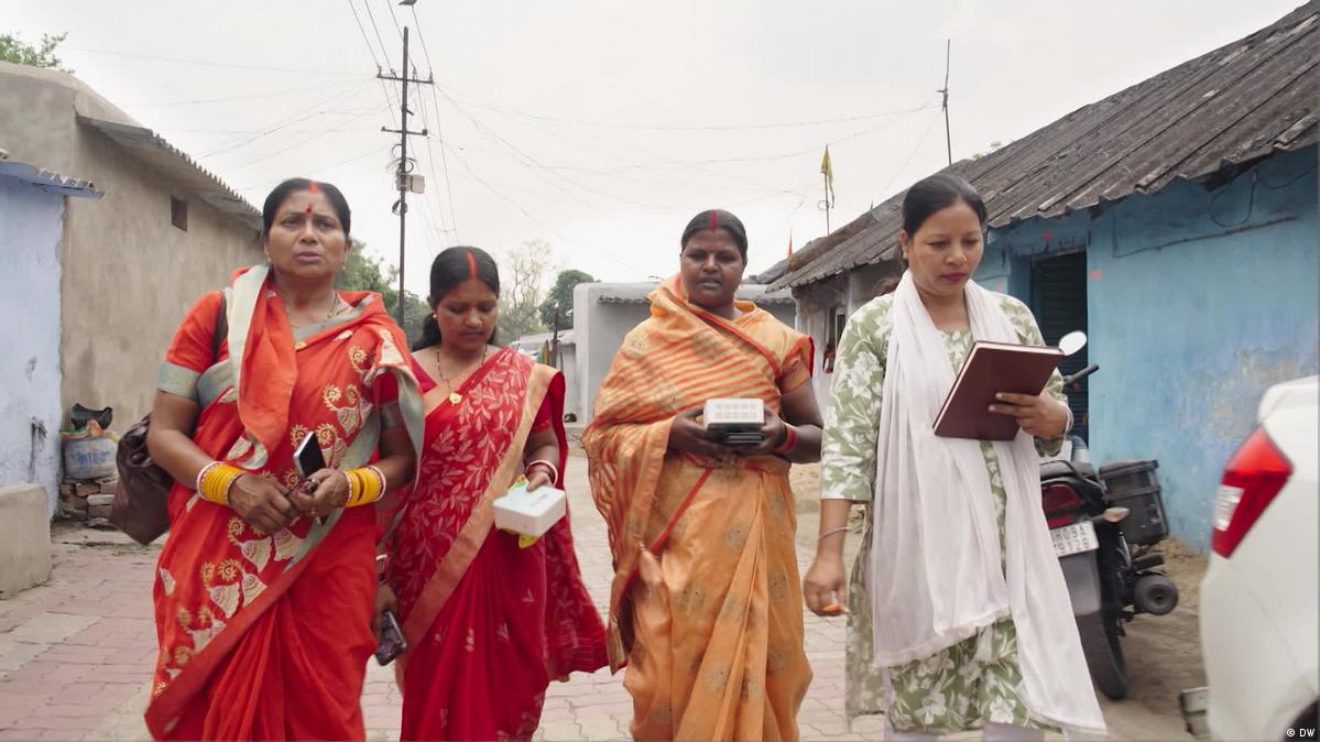 Four women in saris walk through a village street in India, one holding an air-quality monitoring device.