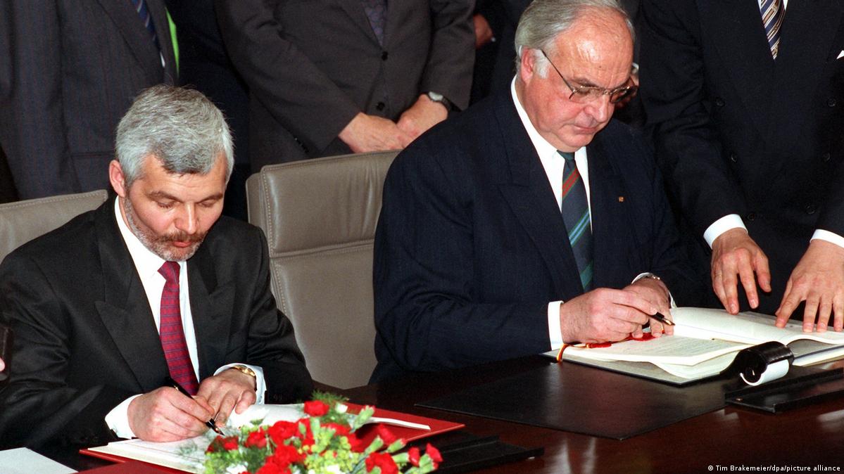 Two men sit beside each other at a desk and sign documents. In the background, officials look on. There are red and white flowers on the table. Bonn, Germany, June 17, 1991