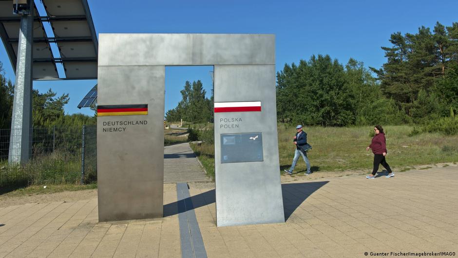 Dva lidé kráčejí směrem k kovovému rámu s otvorem uprostřed. Čára na zemi prochází otvorem. Vlajky a názvy obou zemí (v polštině a němčině) se objevují na rámu na obou stranách otvoru. V pozadí je cesta a stromy. Ahlbeck, Usedom, Německo, 10. června 2025.