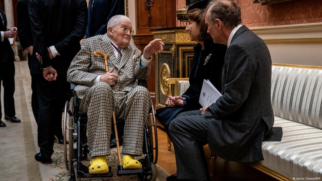 David Hockney en silla de ruedas, con crocs amarillas y bastón, habla con dos personas sentadas en un banco de un palacio. David Hockney en silla de ruedas, con crocs amarillas y bastón, habla con dos personas sentadas en un banco de un palacio.