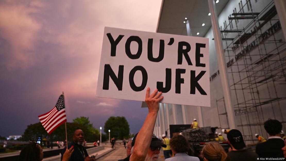 Un hombre sostiene un cartel con la leyenda "Tú no eres JFK", en señal de protesta mientras la gente observa las obras para retirar el nombre de Donald Trump de la señalización del Centro Kennedy. (12.06.2026)