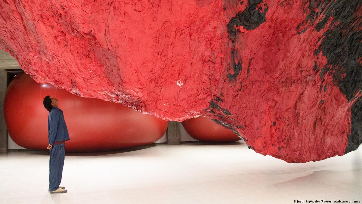 A staff member views 'Mount Moriah at the Gate of the Ghetto' by Anish Kapoor, huge red and black rock-like installation hanging from the ceiling.